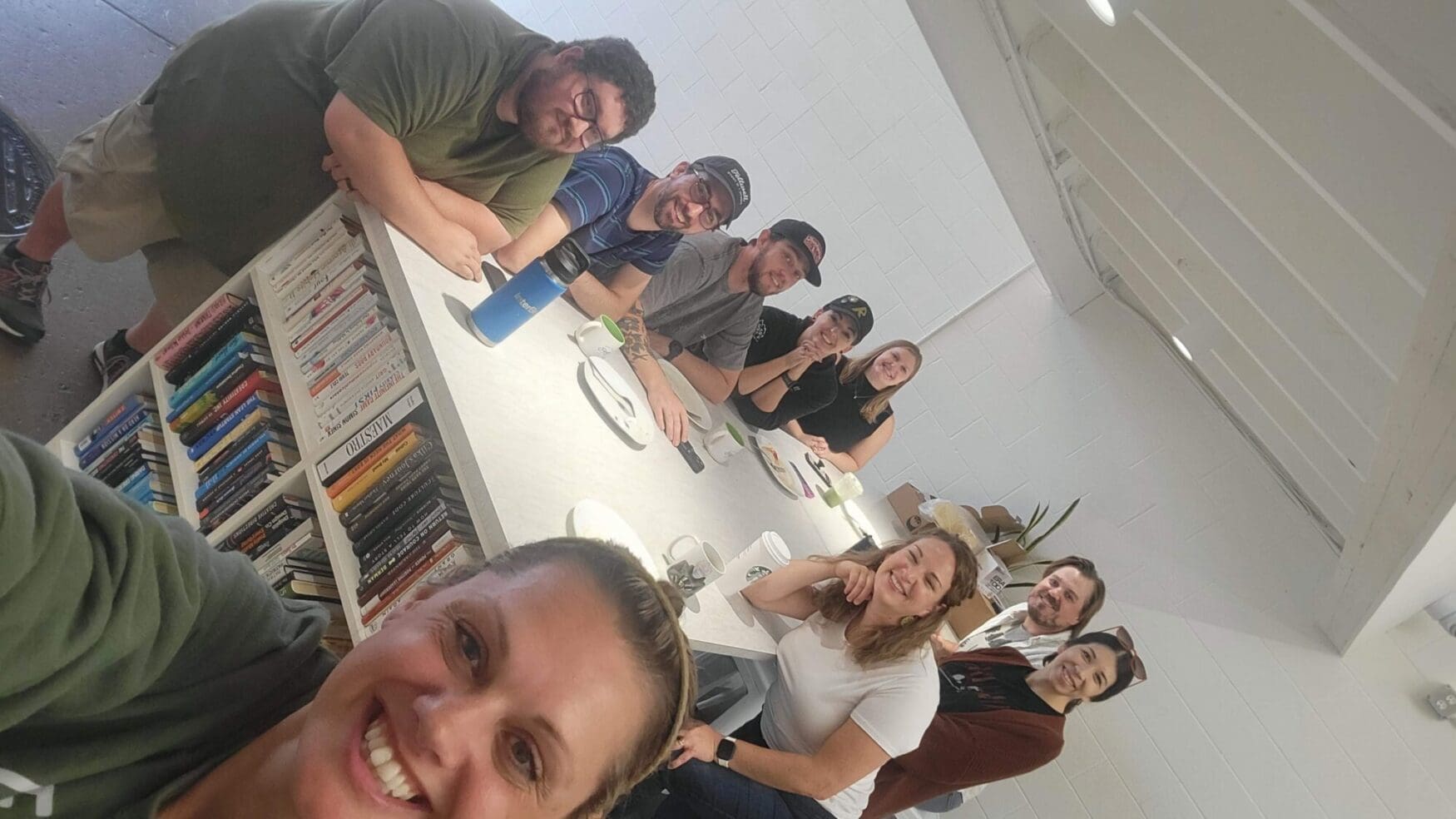 A group of smiling Tellwell team members sitting together around a long white table, mugs and plates in front of them.