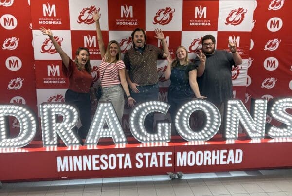 Five people pose enthusiastically in front of a red-and-white Minnesota State University Moorhead step-and-repeat backdrop. They stand behind large illuminated letters spelling “DRAGONS,” with “MINNESOTA STATE MOORHEAD” displayed below.
