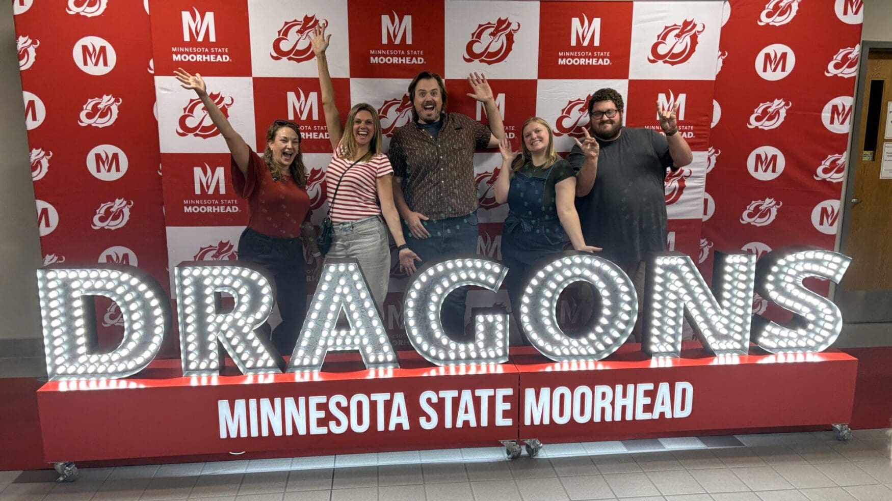 Five people pose enthusiastically in front of a red-and-white Minnesota State University Moorhead step-and-repeat backdrop. They stand behind large illuminated letters spelling &ldquo;DRAGONS,&rdquo; with &ldquo;MINNESOTA STATE MOORHEAD&rdquo; displayed below.