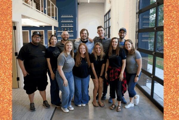 A group of eleven smiling people stand together in a modern office with white walls, a staircase, and a glass garage-style door. Everyone is casually dressed, and they pose closely as a team.