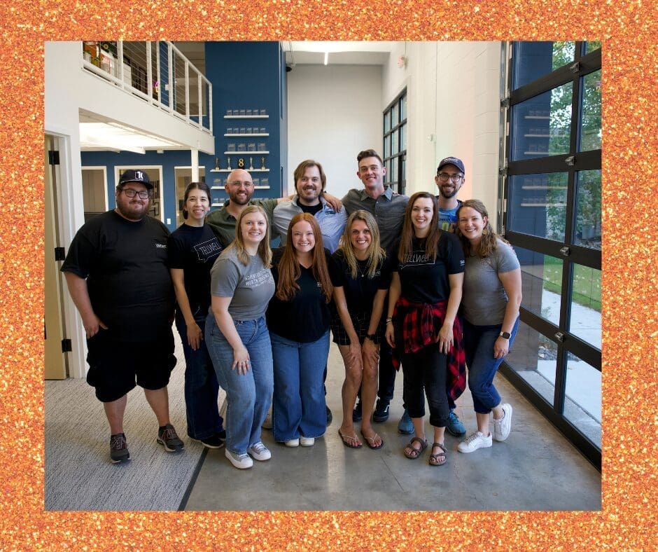 A group of eleven smiling people stand together in a modern office with white walls, a staircase, and a glass garage-style door. Everyone is casually dressed, and they pose closely as a team.