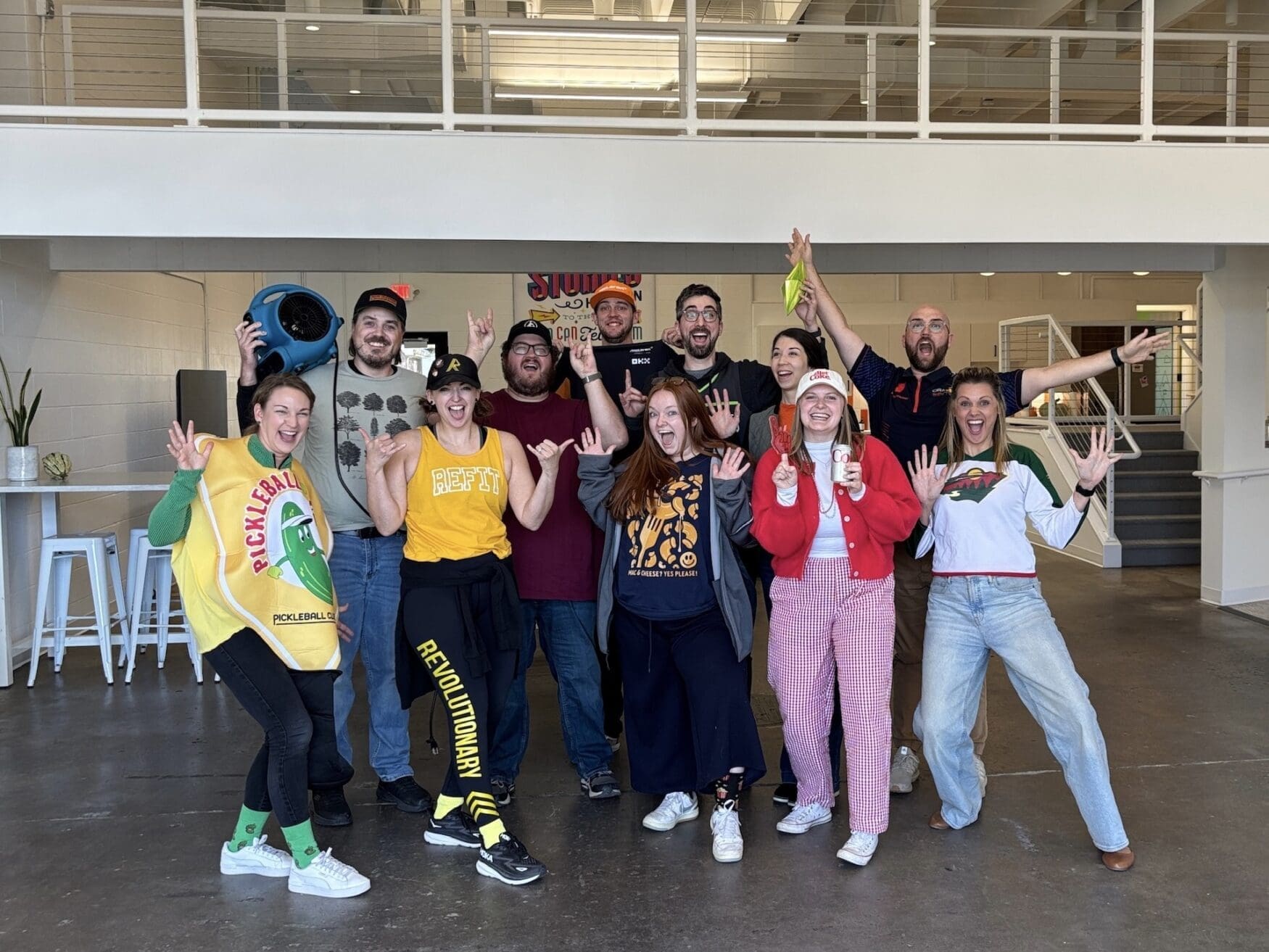 A group of 11 men and women pose for a group photo. They all have excited facial expressions and are wearing festive costumes. They are in a large office space with an indoor walkway behind them.