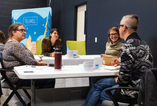 A group of four people seated around a white table. They are in a room with black walls with a backdrop behind them that reads “Tellwell”. They have notebooks and additional work objects on the table and are having a discussion.
