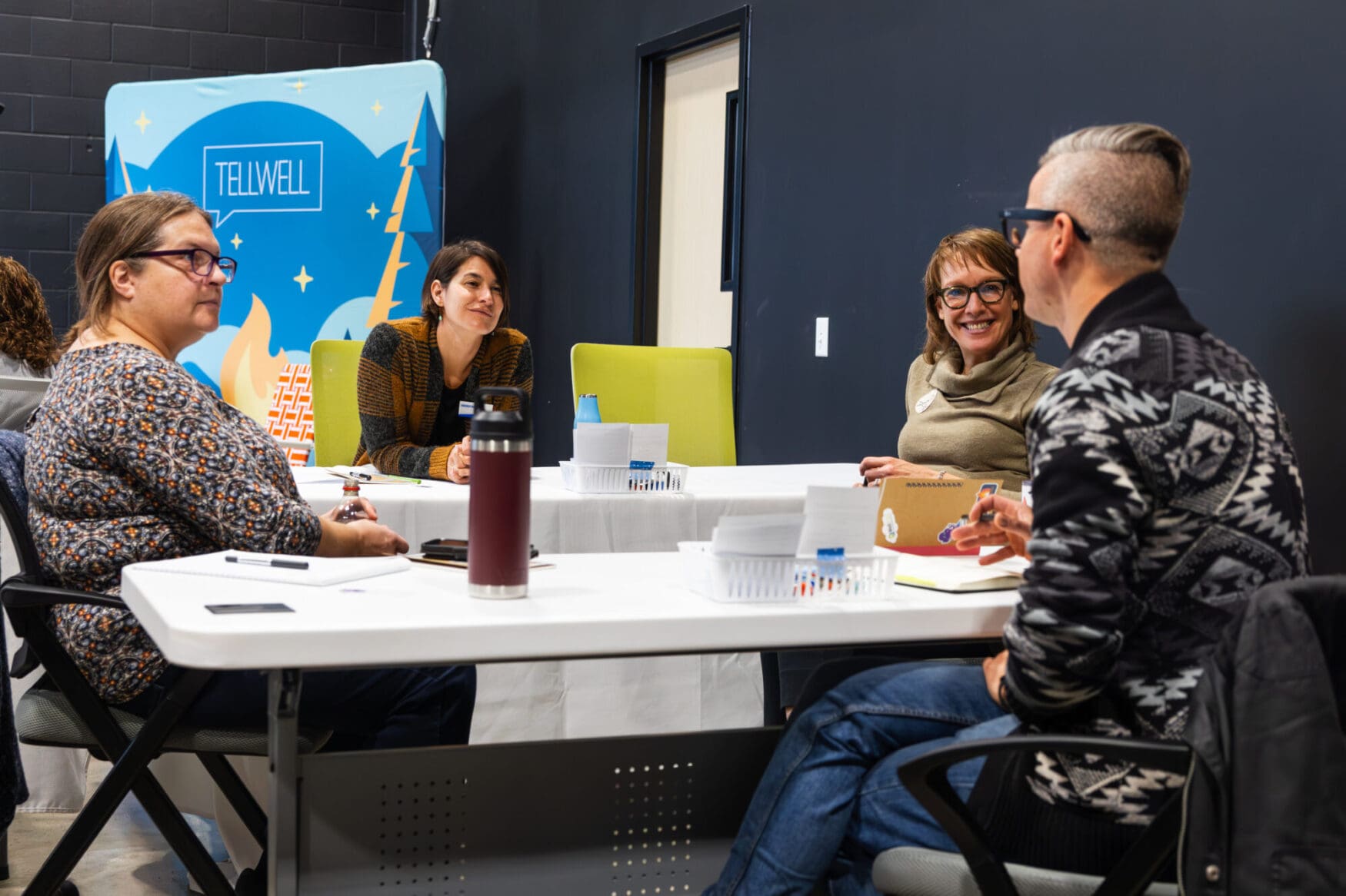 A group of four people seated around a white table. They are in a room with black walls with a backdrop behind them that reads &ldquo;Tellwell&rdquo;. They have notebooks and additional work objects on the table and are having a discussion.