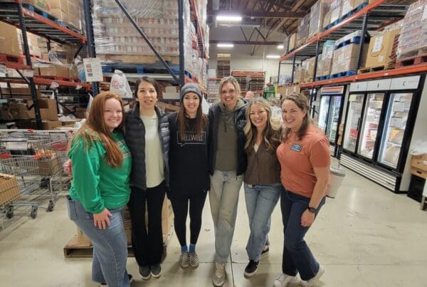 A group of six women stand for a photo inside a warehouse. They are in a food pantry and appear glad to be volunteering.