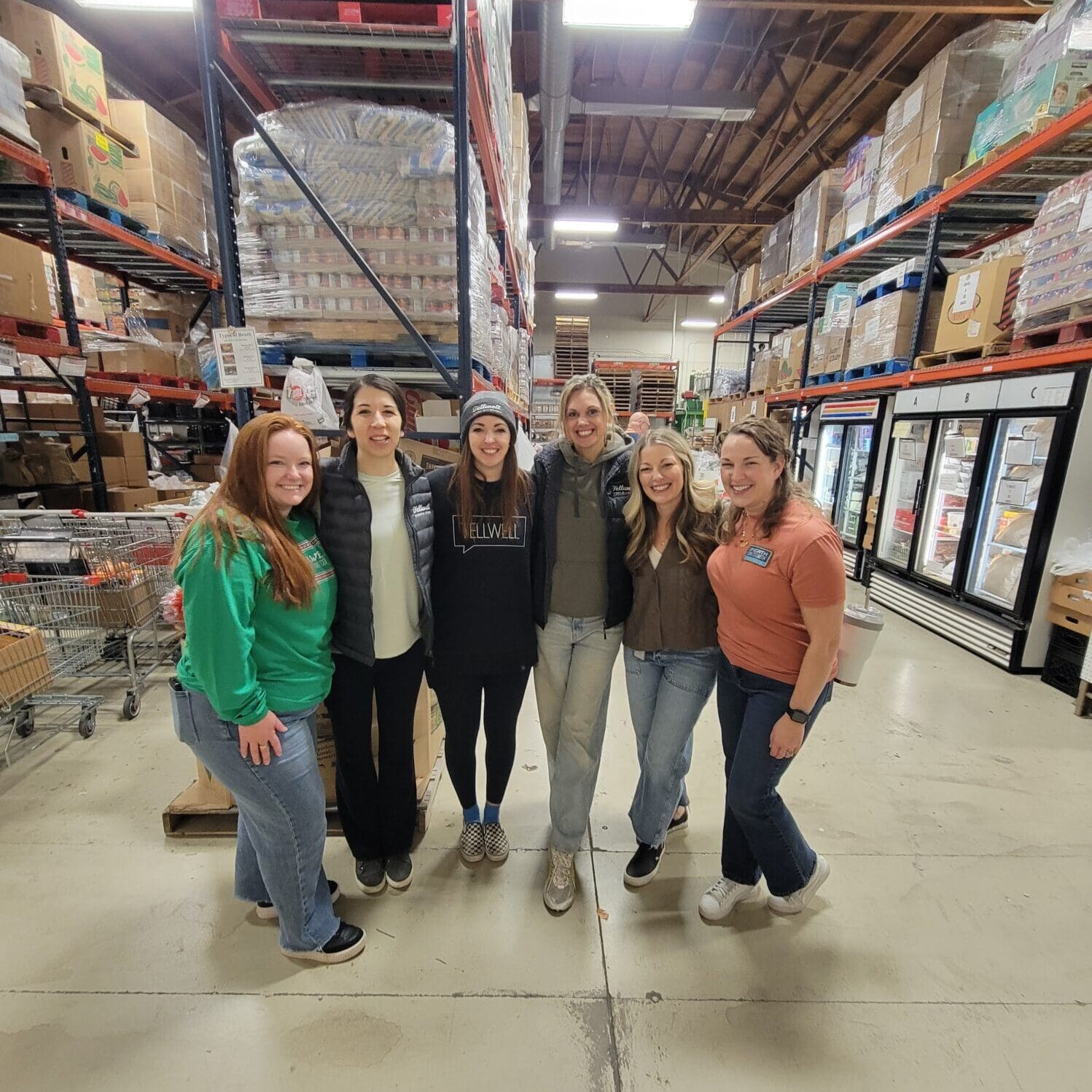 A group of six women stand for a photo inside a warehouse. They are in a food pantry and appear glad to be volunteering.