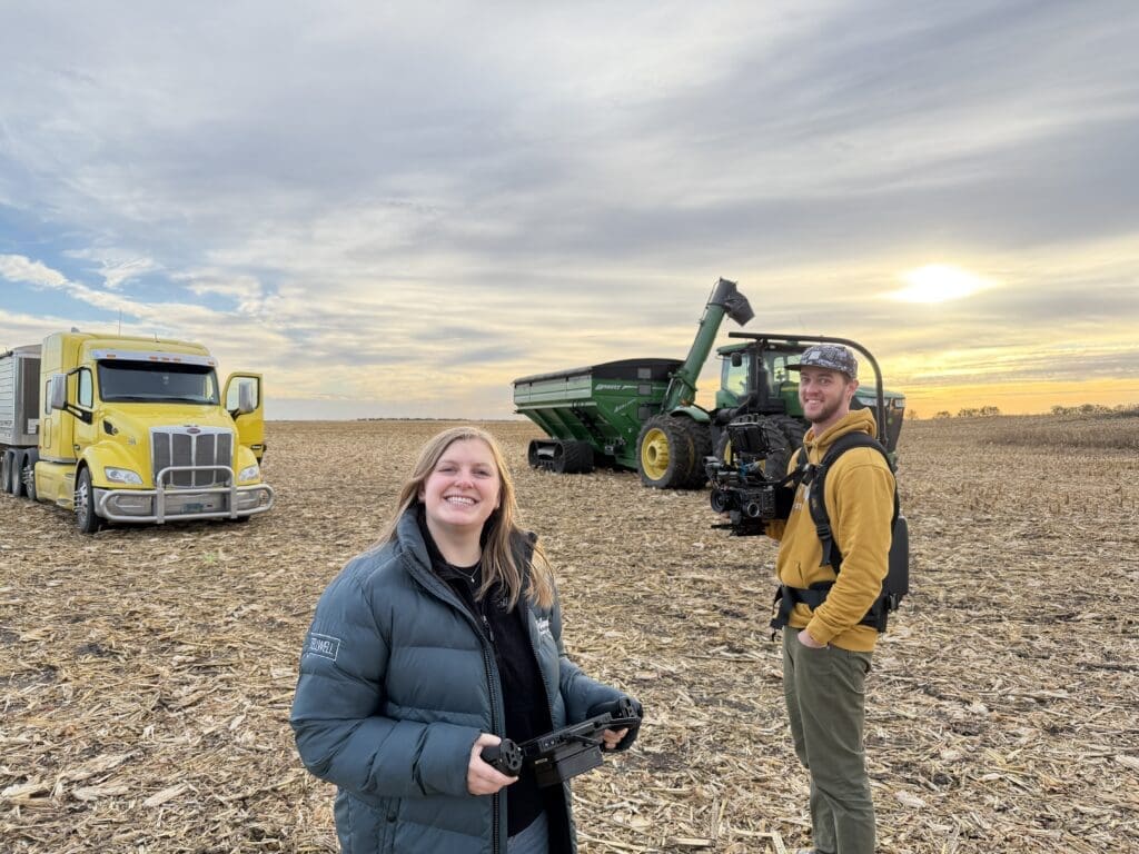Two people standing in a harvested cornfield at sunrise, one holding a camera monitor and the other carrying a camera rig. A yellow semi-truck and green tractor are parked nearby under a partly cloudy sky.