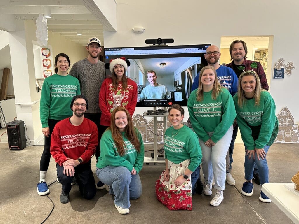 Team members wearing festive Tellwell sweaters pose together in an office during a holiday celebration, smiling in front of a screen showing a remote teammate on a video call.