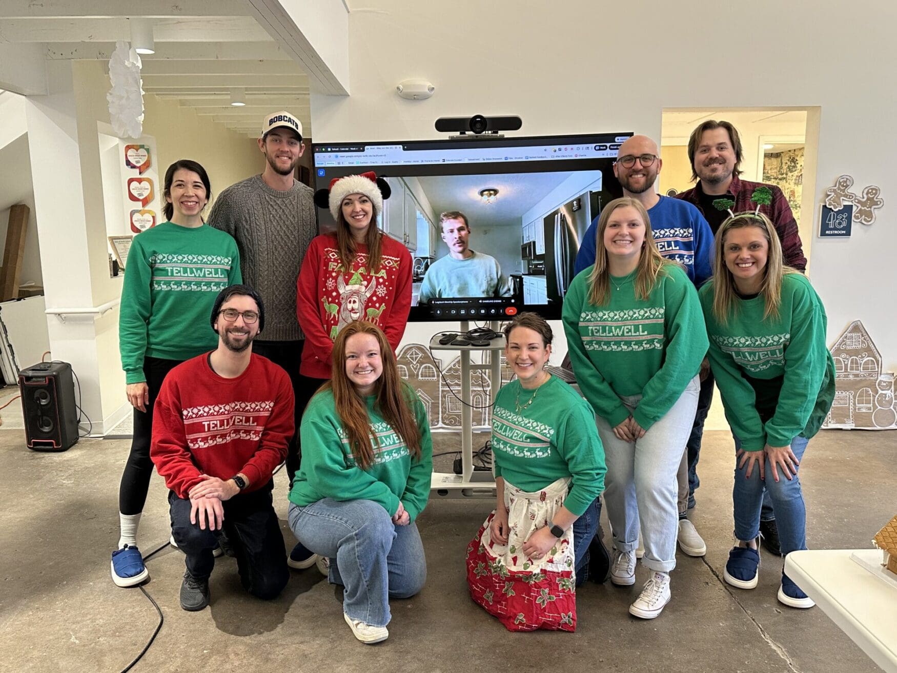 Team members wearing festive Tellwell sweaters pose together in an office during a holiday celebration, smiling in front of a screen showing a remote teammate on a video call.