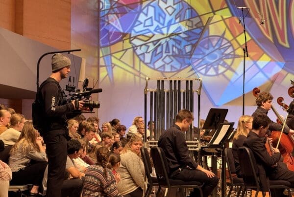 A cameraman in a beanie films an orchestra from the side as colorful stained glass designs are projected on the wall. The audience watches intently.