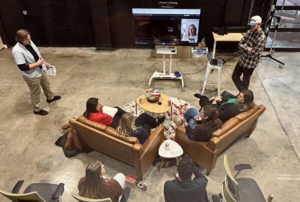 A high-angle shot of an informal team meeting in a large industrial-style studio space. Five colleagues sit on leather sofas and chairs in a circle around a coffee table with snacks, while two others stand nearby. A large television monitor in the background shows a remote participant on a video call.