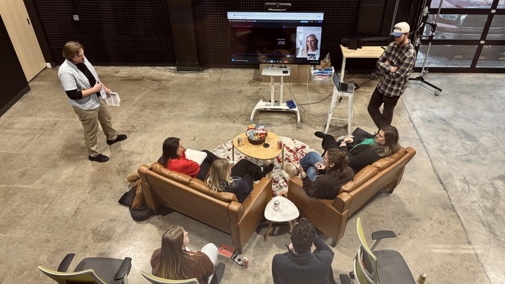 A high-angle shot of an informal team meeting in a large industrial-style studio space. Five colleagues sit on leather sofas and chairs in a circle around a coffee table with snacks, while two others stand nearby. A large television monitor in the background shows a remote participant on a video call.