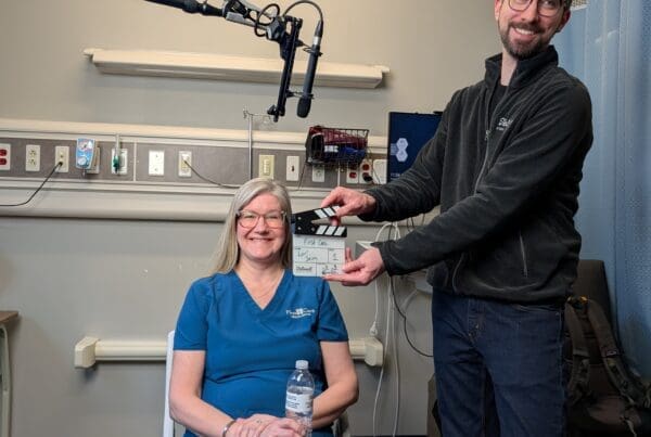 A woman in blue medical scrubs sits in a hospital room holding a water bottle, smiling at the camera. A man in a black fleece jacket stands next to her, holding a film slate that reads "First Care." A professional boom microphone hangs from the ceiling above them.