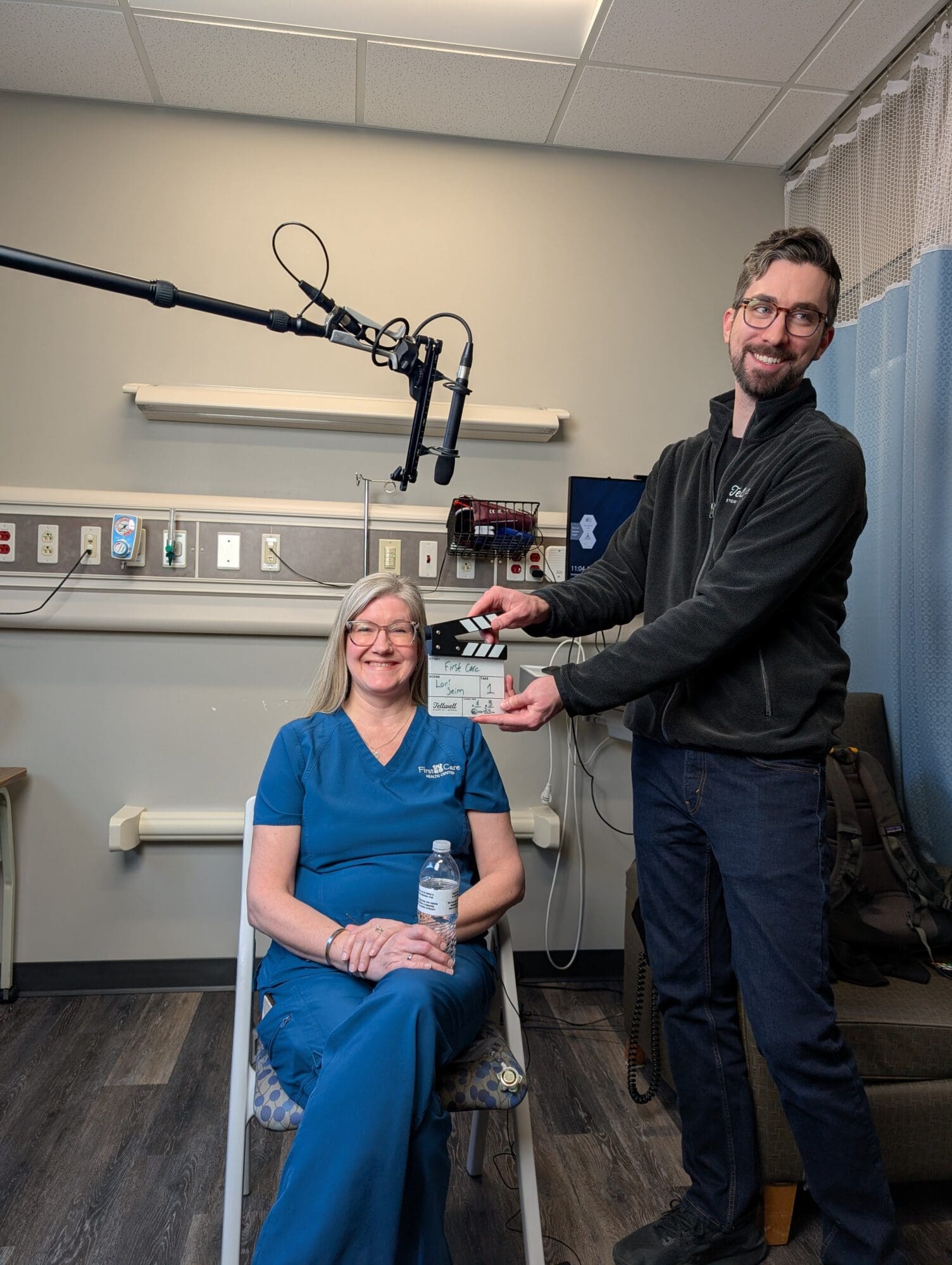 A woman in blue medical scrubs sits in a hospital room holding a water bottle, smiling at the camera. A man in a black fleece jacket stands next to her, holding a film slate that reads "First Care." A professional boom microphone hangs from the ceiling above them.