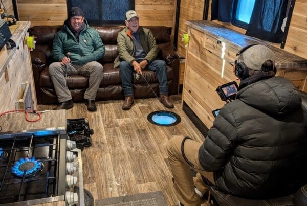 Inside a wood-paneled ice fishing house, a videographer in a black puffer jacket films two men sitting on a leather sofa. The men are smiling and holding fishing rods over a glowing blue hole in the floor, while a gas stove burner glows blue in the foreground.