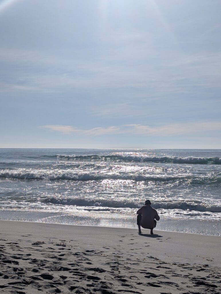 A person in a light-colored shirt crouches on a sandy beach, looking out at the shimmering ocean waves. The sun reflects brightly off the water's surface under a clear, pale blue sky.
