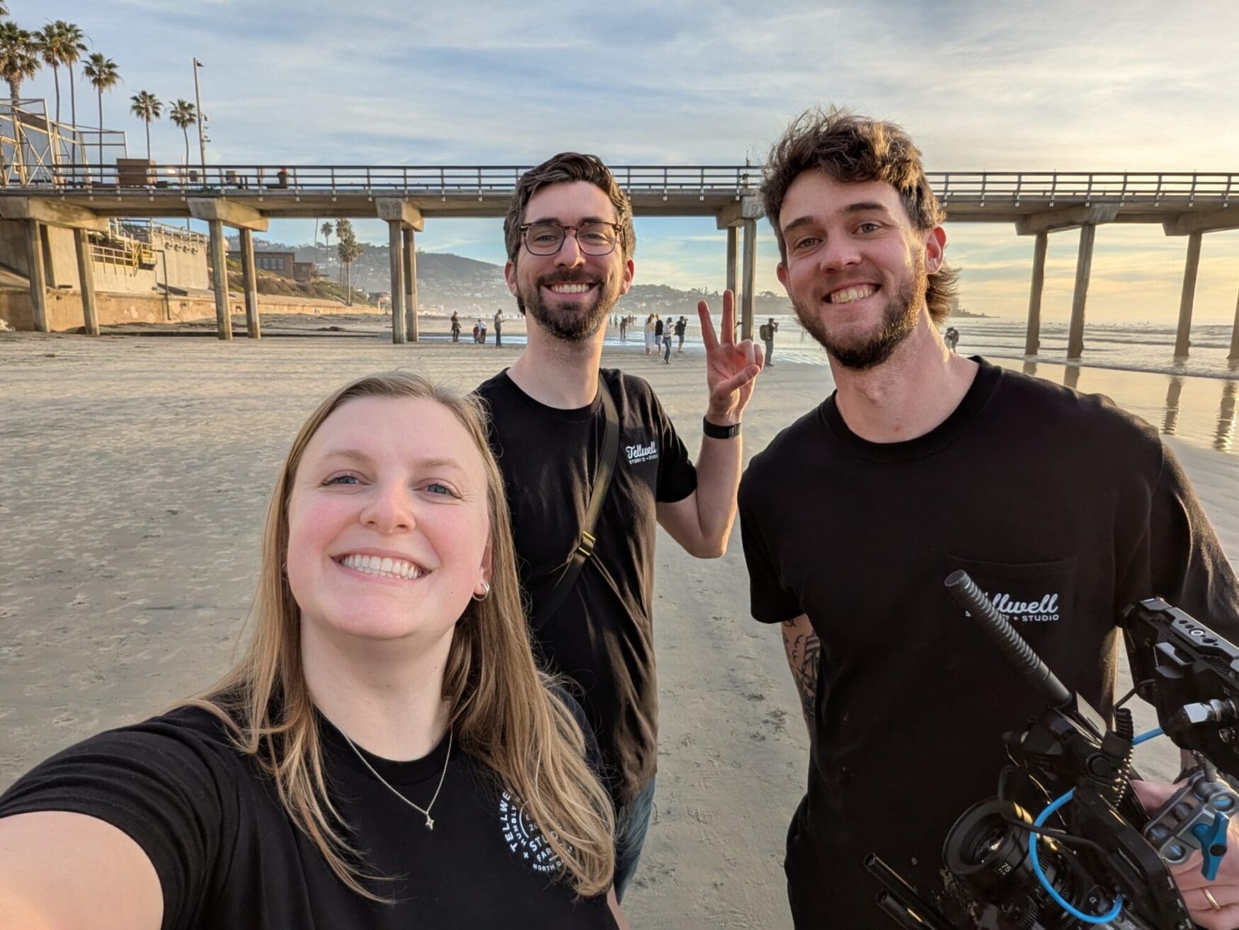Three coworkers smile for a selfie on a wide, sandy beach during the "golden hour." A long wooden pier stretches into the ocean behind them, and one man holds a professional video camera rig.