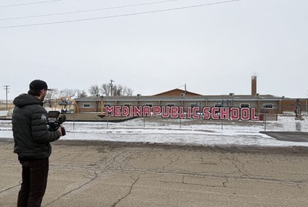 A videographer in a puffer jacket films the exterior of Medina Public School. The school's name is displayed in large, red and white block letters on a fence in front of the low-profile brick building on a gray, overcast day.