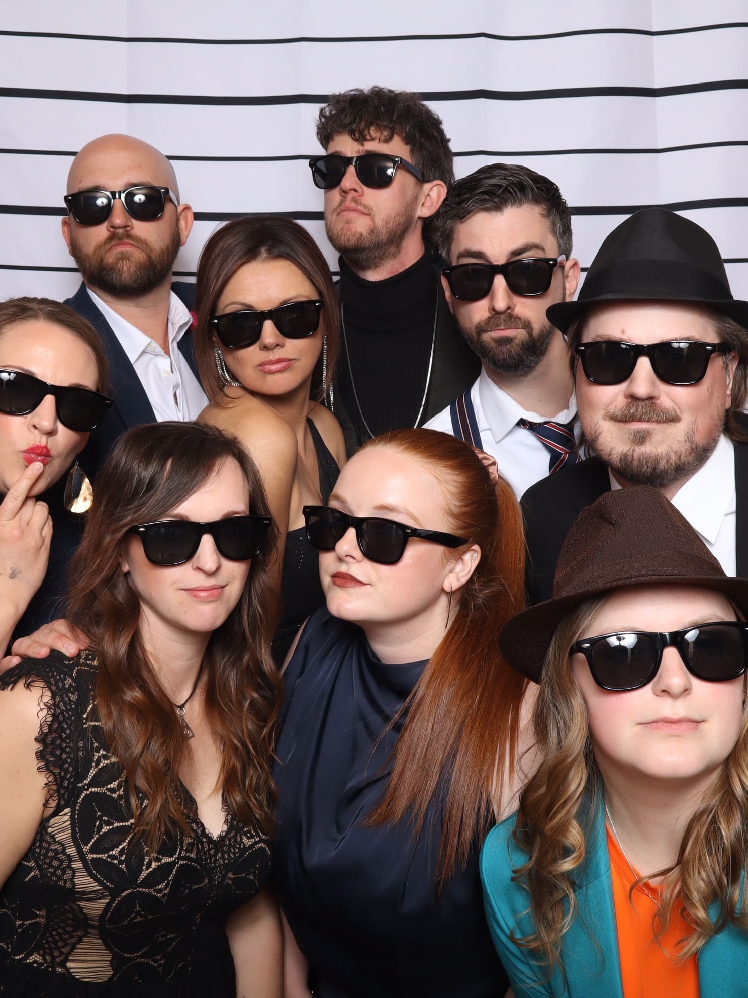 A group photo of nine people posing with serious expressions and wearing matching black sunglasses. They are standing in front of a white backdrop with horizontal black lines, mimicking a police lineup.