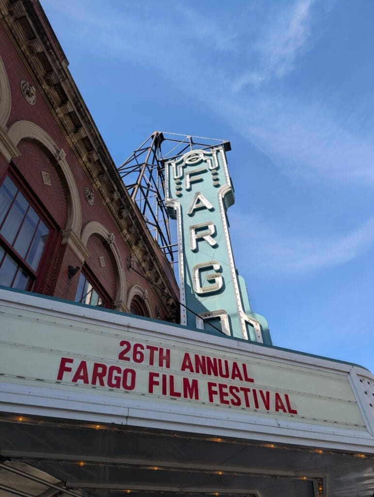 A low-angle shot of the Fargo Theatre&rsquo;s exterior, showcasing the red brick architecture and the teal-colored vintage neon sign that spells out "FARGO." The marquee below displays "26TH ANNUAL FARGO FILM FESTIVAL" in bold red lettering.