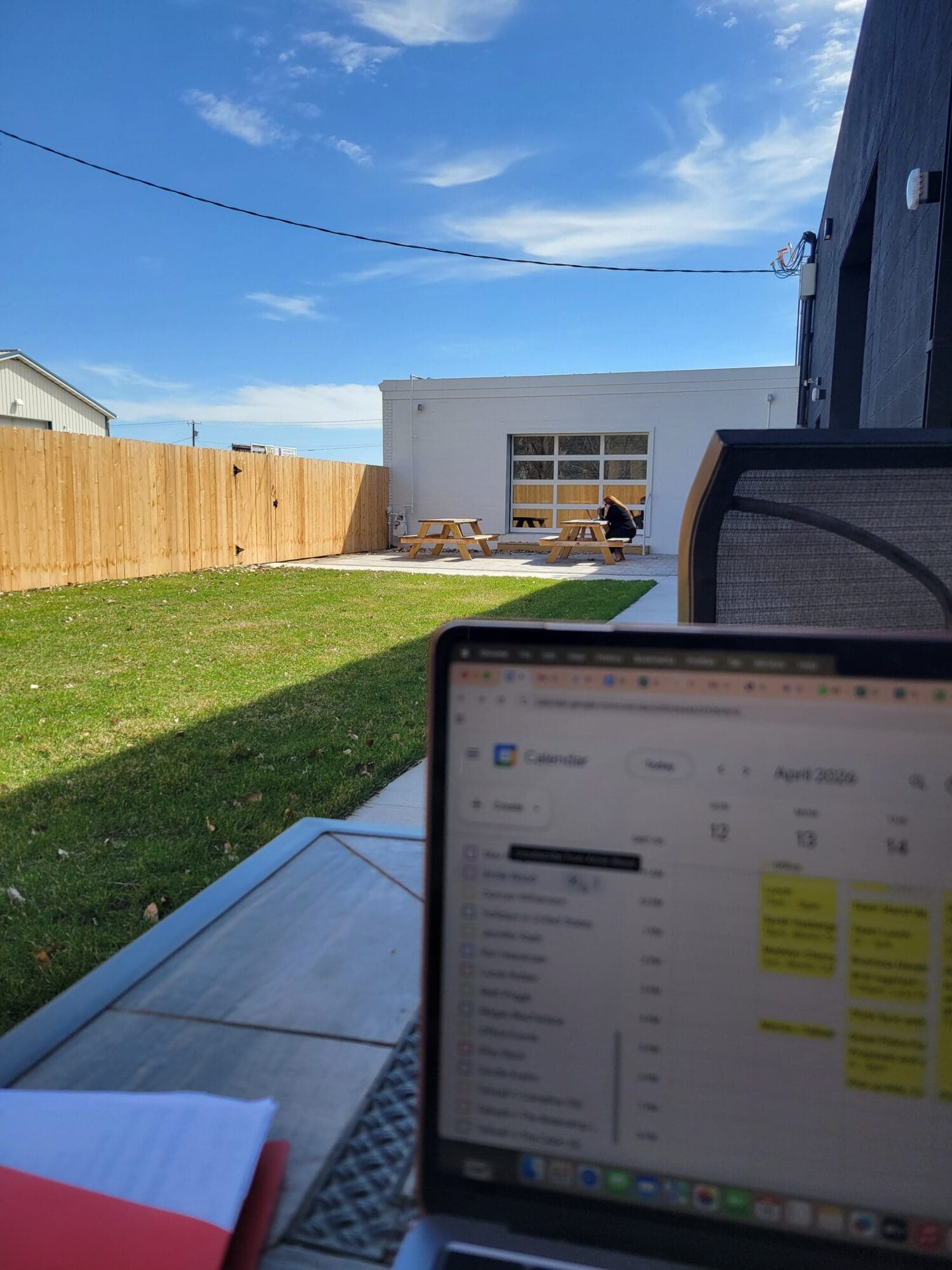 A first-person view from an outdoor table showing a laptop screen with a Google Calendar. In the background, a sunny patio area features a green lawn, a wooden fence, and a person working at a picnic table under a clear blue sky.