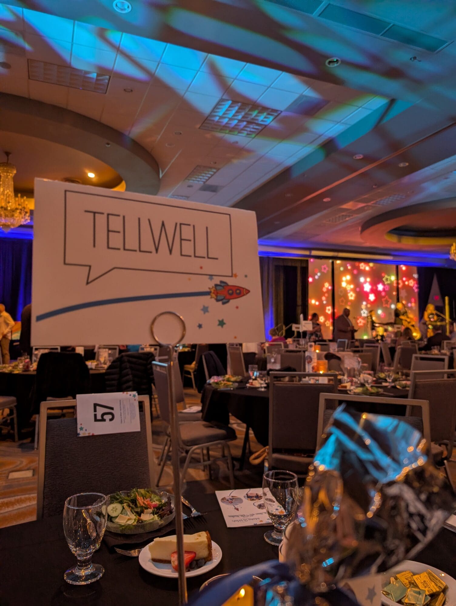 A table at a formal event features a place card with the "Tellwell" logo and a rocket ship illustration. In the background, a large ballroom is filled with guests at round tables under colorful stage lighting and a crystal chandelier.