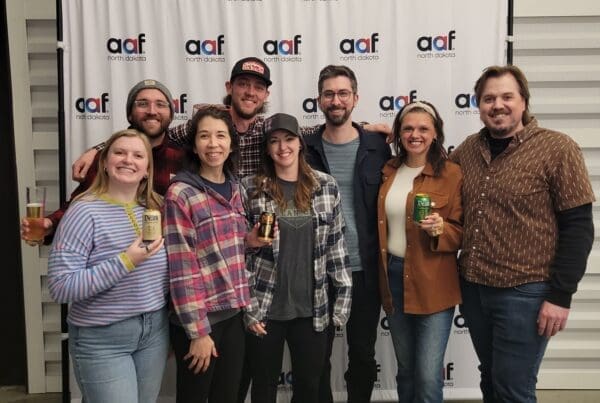Seven people stand in a group in front of an "AAF North Dakota" branded backdrop. Several team members are smiling and holding up cans of Liquid Death sparkling water and glasses of beer.