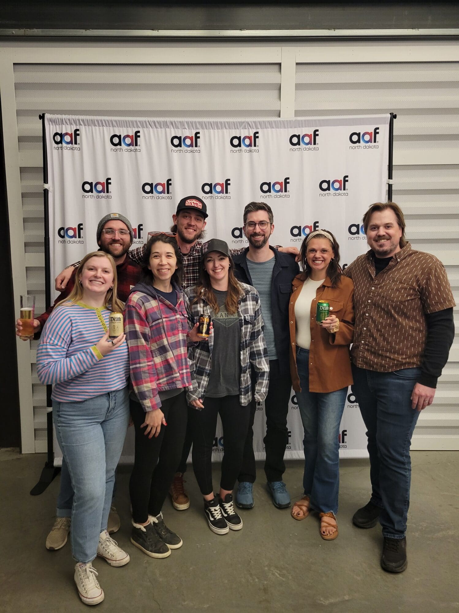 Seven people stand in a group in front of an "AAF North Dakota" branded backdrop. Several team members are smiling and holding up cans of Liquid Death sparkling water and glasses of beer.