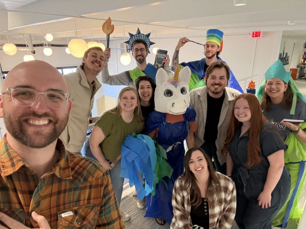 A cheerful group photo of the team in a modern office hallway. Several members are wearing the DIY Statue of Liberty and unicorn costumes made from colorful plastic and tissue paper, while others lean in to join the shot. A man in a plaid shirt takes the selfie in the foreground, capturing everyone&rsquo;s high energy and smiles.