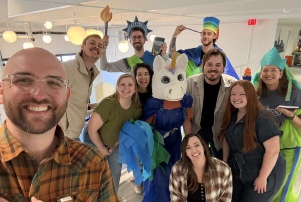 A cheerful group photo of the team in a modern office hallway. Several members are wearing the DIY Statue of Liberty and unicorn costumes made from colorful plastic and tissue paper, while others lean in to join the shot. A man in a plaid shirt takes the selfie in the foreground, capturing everyone&rsquo;s high energy and smiles.