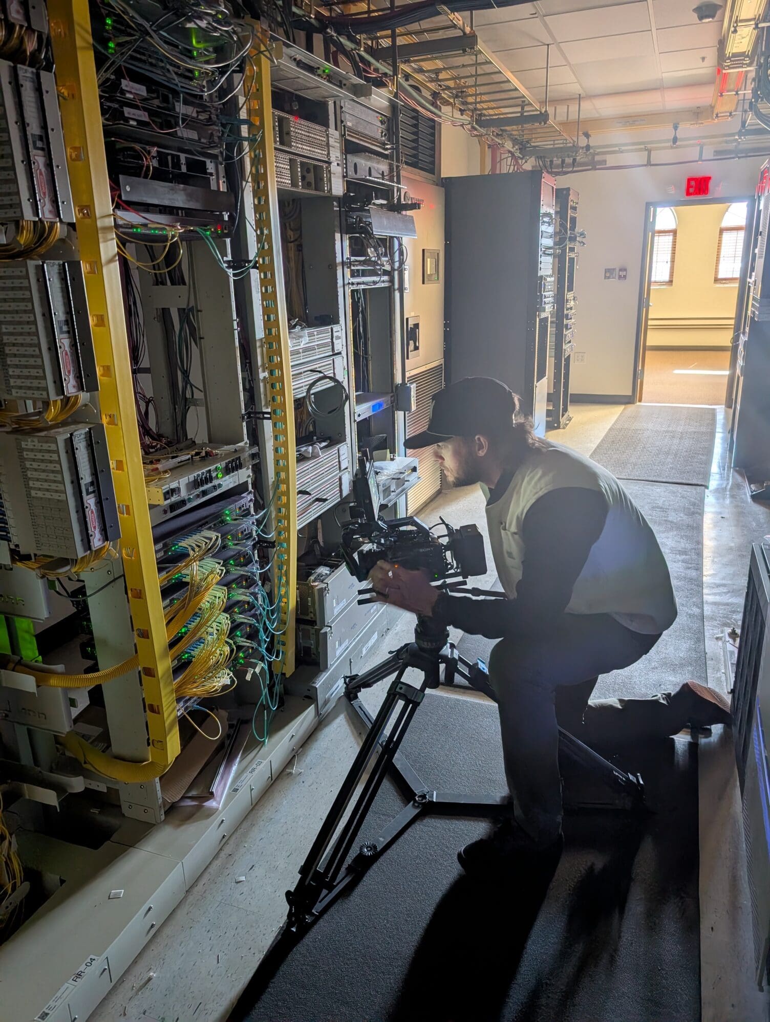 A videographer wearing a baseball cap and safety vest kneels on a walkway, using a tripod-mounted camera to film a complex wall of network server racks. The server room is filled with intricate wiring and glowing indicator lights.